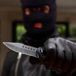 Burglar with balaclava, holding a knife in left hand, armed robbery concept. Robber threaten with a dagger, blur indoor background, closeup view.