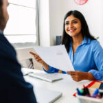 Confident successful Indian female HR recruitment manager holding resume in hands while having an interview in a modern office, looking and listening to job candidate. Friendly interview between HR female director and caucasian job candidate.
