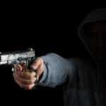 Gun in male human hand aiming toward left side in dark. The person is hidden in shadow, only hand and gun is illuminated. Shot indoor with a full frame DSLR camera.